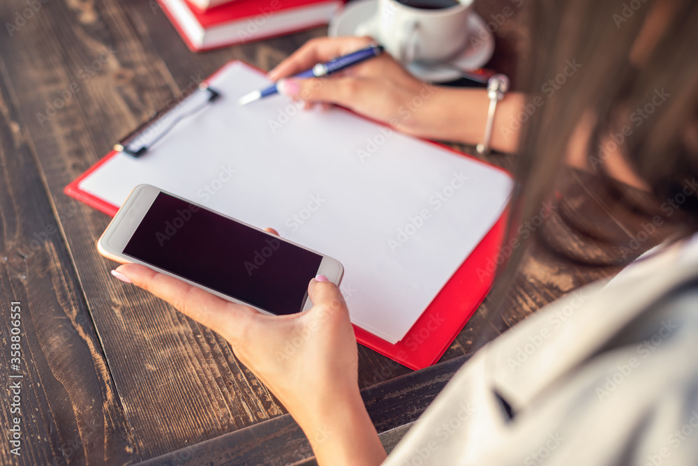 Mockup image of a woman's hands holding white mobile phone with blank black screen on background of white sheet of paper at a cafe. Mockup image of a woman's hands holding white mobile phone with blank black screen on background of white sheet of paper at a cafe.