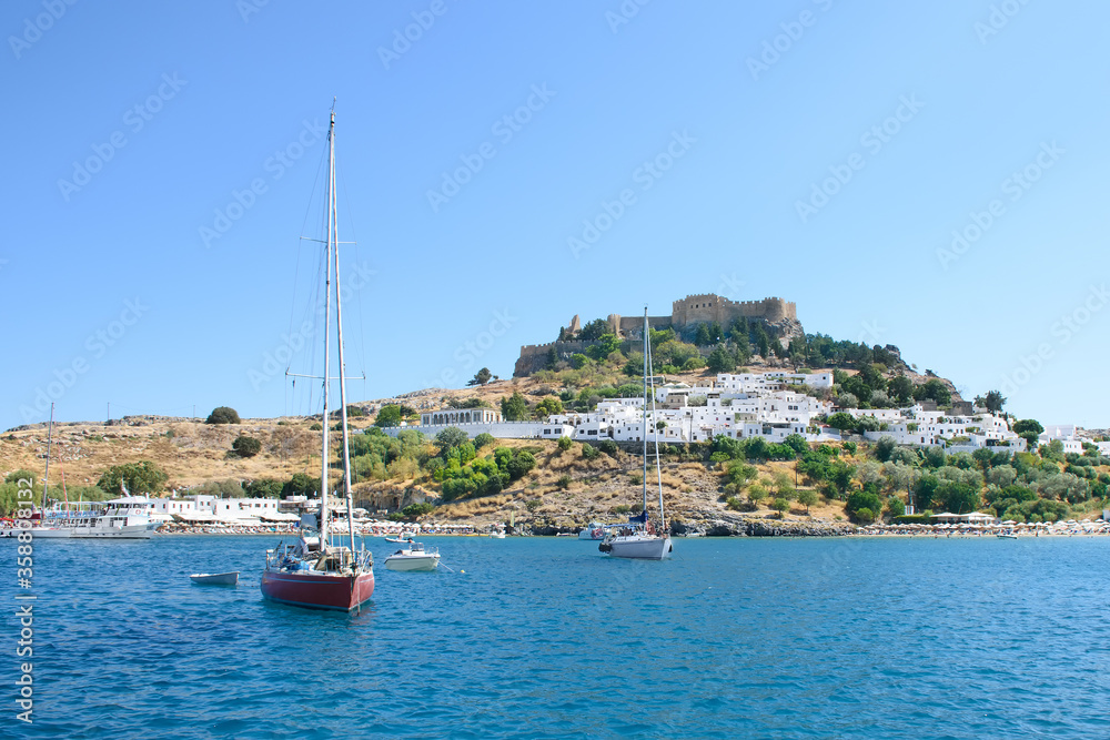 Sailing boats anchored in bay right under Village of Lindos with Acropolis on hill (Rhodes, Greece)