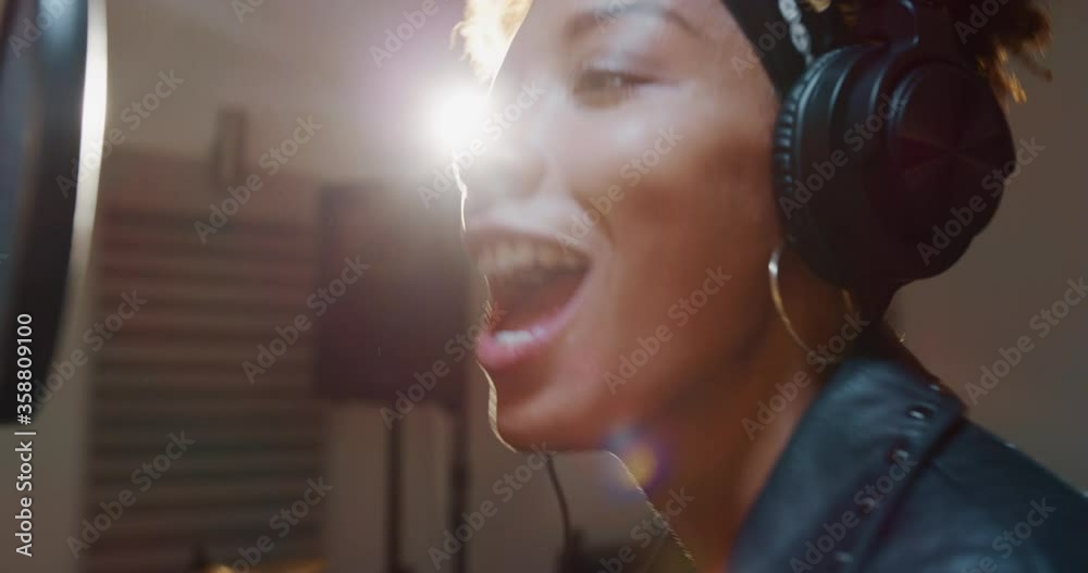 Close up of an young professional smiling energetic african female singer wearing bandana and headphones is performing a new song with a microphone while recording it in a music studio. 