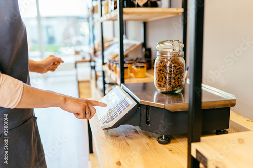 Female shop owner weighs glass jar with dark brown whole grains of pasta. Woman assistant in zero waste shop. Weighing dry goods in plastic free grocery store. Sustainable shopping at local business