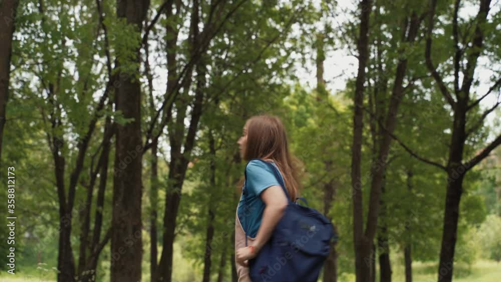 Beautiful woman smiles walking along path surrounded by foliage and trees. Back view of female with backpack walking at the summer forest. Beautiful woman smiles walking along path surrounded by foliage and trees. Back view of female with backpack walking at the summer forest.