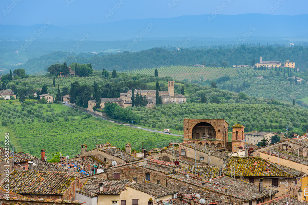 Rooftops of San Gimignano, Tuscan countryside with rolling hills and Santa Maria Assunta a Monte Oliveto Minore church in the distance seen from Parco della Rocca, San Gimignano.