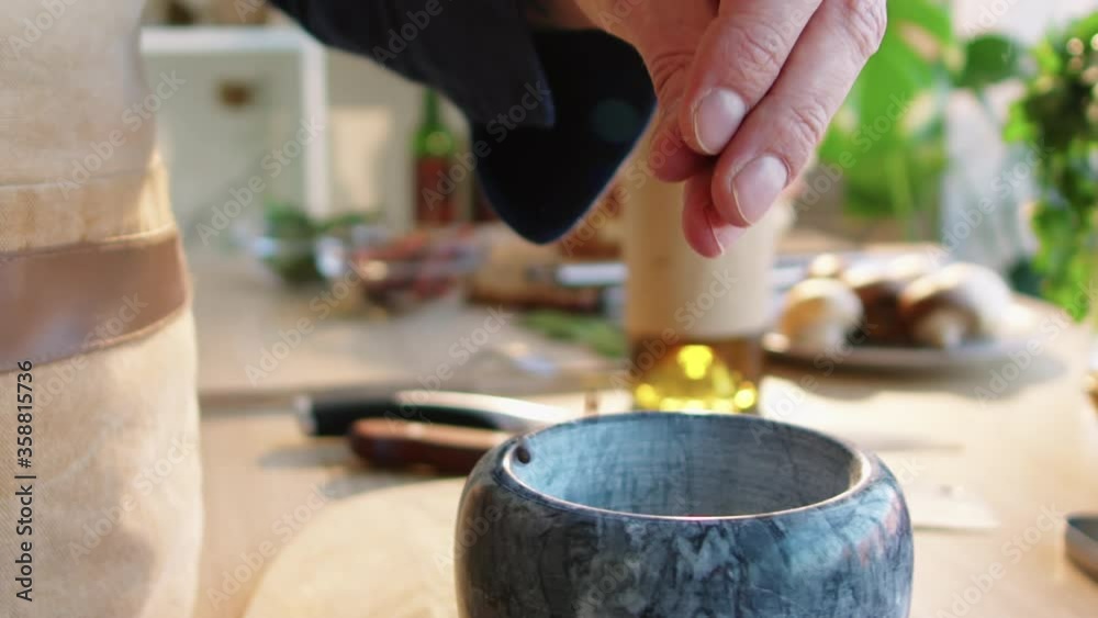 Close up view of hands of unrecognizable male chef picking up pinch of black peppercorns, pouring them into stone mortar and grinding with pestle at kitchen table Close up view of hands of unrecognizable male chef picking up pinch of black peppercorns, pouring them into stone mortar and grinding with pestle at kitchen table
