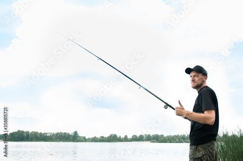 A fisherman in a black T-shirt, cap and camouflage shorts stands on the river bank with a classic fishing rod, free space for text