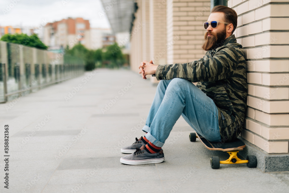 Young bearded man sitting on his skateboard in a skate park on a sunny day.