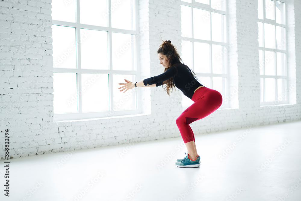 Young girl dressed in trendy outfit for training doing squatting exercises tensing butt muscles working on perfect lower body shape, determined woman going in for sport to reach fitness goals