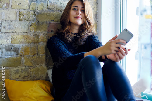 Half length portrait of charming confident teenager looking at camera while using modern technology for communicating sitting in cafe near to background with copy space for your advertising content photo