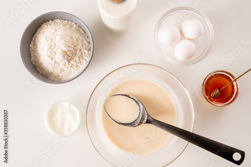 Overview of glass bowls with eggs, honey, dough for pancakes, sourcream, flour