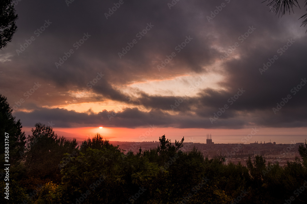 dramatic sunrise view of barcelona at dusk from guinardo park, with sun rising through clouds and cityscape with tres xemeneies