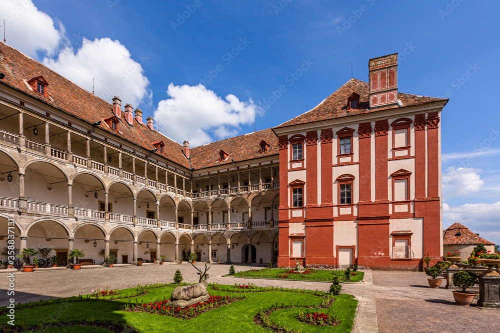 Opocno / Czech Republic - June 16 2020: View of the castle courtyard with arcades and red facade. Green lawn with statue and flowers in foreground. Sunny summer day with blue sky and white clouds.