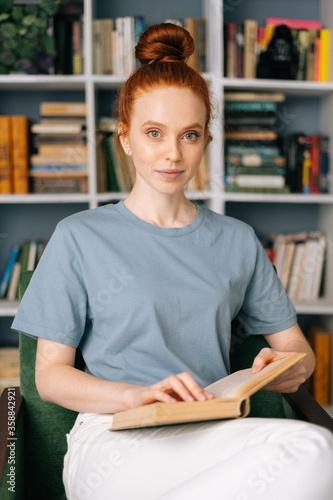 Loving redhead young woman student is reading book enjoys of rest at home office, looking at camera. Cute lady enjoying books at library room on background of bookshelves.