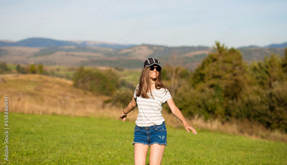 Carefree happy woman in sunglasses, cap and jeans shorts walk on green grass meadow on top of mountain enjoying nature. Freedom Carefree happy woman in sunglasses, cap and jeans shorts walk on green grass meadow on top of mountain enjoying nature. Freedom