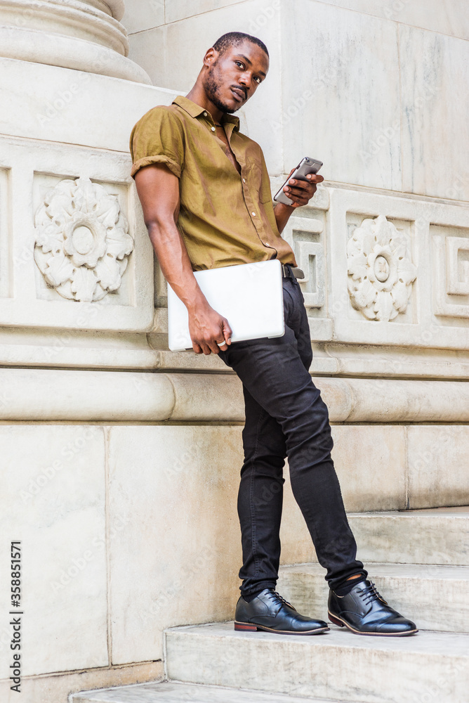Young African American Man studying in New York, wearing green short sleeve shirt, black pants, leather shoes, carrying laptop computer, standing against white marble wall, texting on cell phone.. Young African American Man studying in New York, wearing green short sleeve shirt, black pants, leather shoes, carrying laptop computer, standing against white marble wall, texting on cell phone..