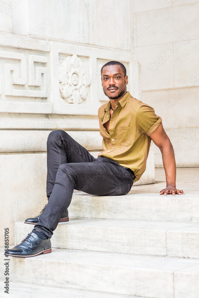 Young African American Male College Student with beard, wearing green short sleeve shirt, black pants, leather shoes, sitting on stairs of office building on campus in New York City, relaxing..