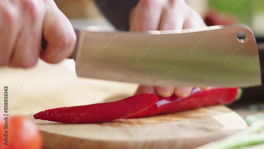 Close-up shot of male hands rolling red chili pepper on wooden board and cutting it into two pieces with knife