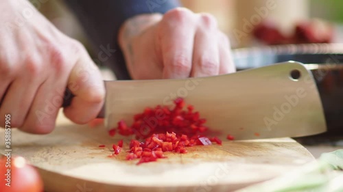 Close-up view of hands of male chef dicing red pepper on wooden cutting board with butcher knife