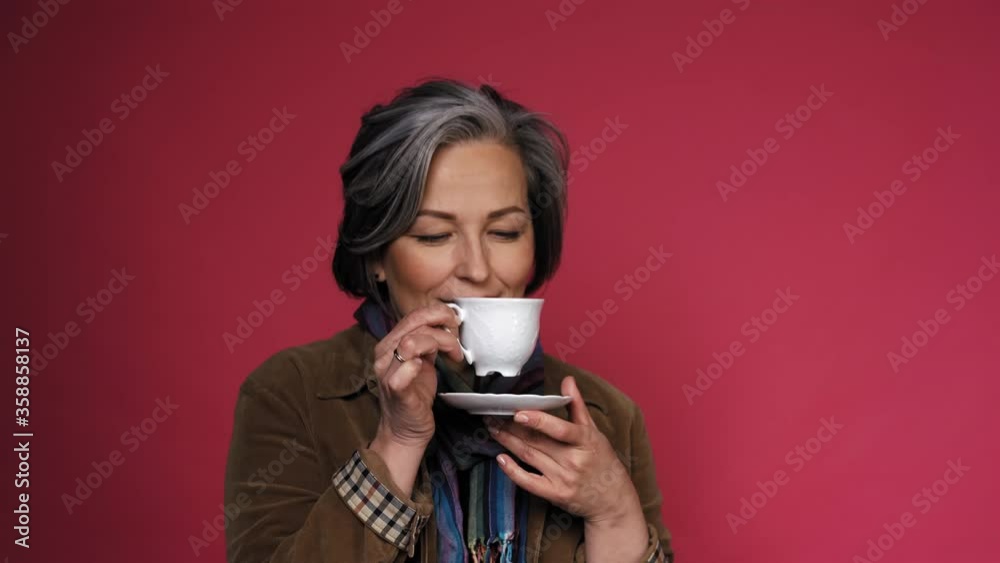 Charming mature woman enjoys cup of coffee with eyes closed. Happy gray-haired lady drinks coffee posing on pink background in the studio. Close up portrait with text space on right. Prores 422. Charming mature woman enjoys cup of coffee with eyes closed. Happy gray-haired lady drinks coffee posing on pink background in the studio. Close up portrait with text space on right. Prores 422.