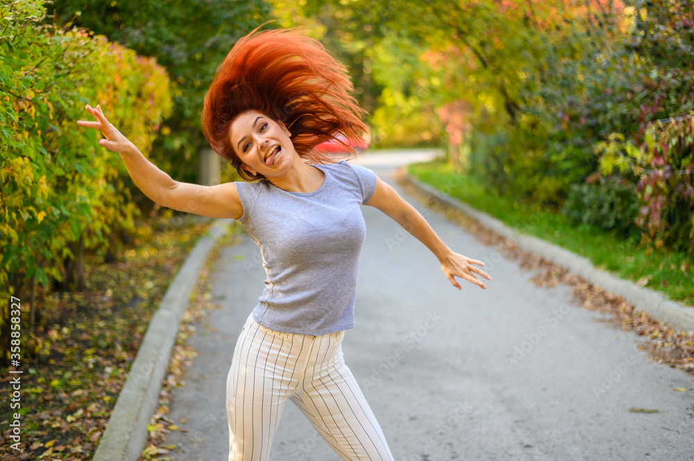 A beautiful red-haired woman is walking along the street and dancing. Happy girl in a gray T-shirt and striped leggings walks and has fun in the autumn park. Photo in motion. A beautiful red-haired woman is walking along the street and dancing. Happy girl in a gray T-shirt and striped leggings walks and has fun in the autumn park. Photo in motion.