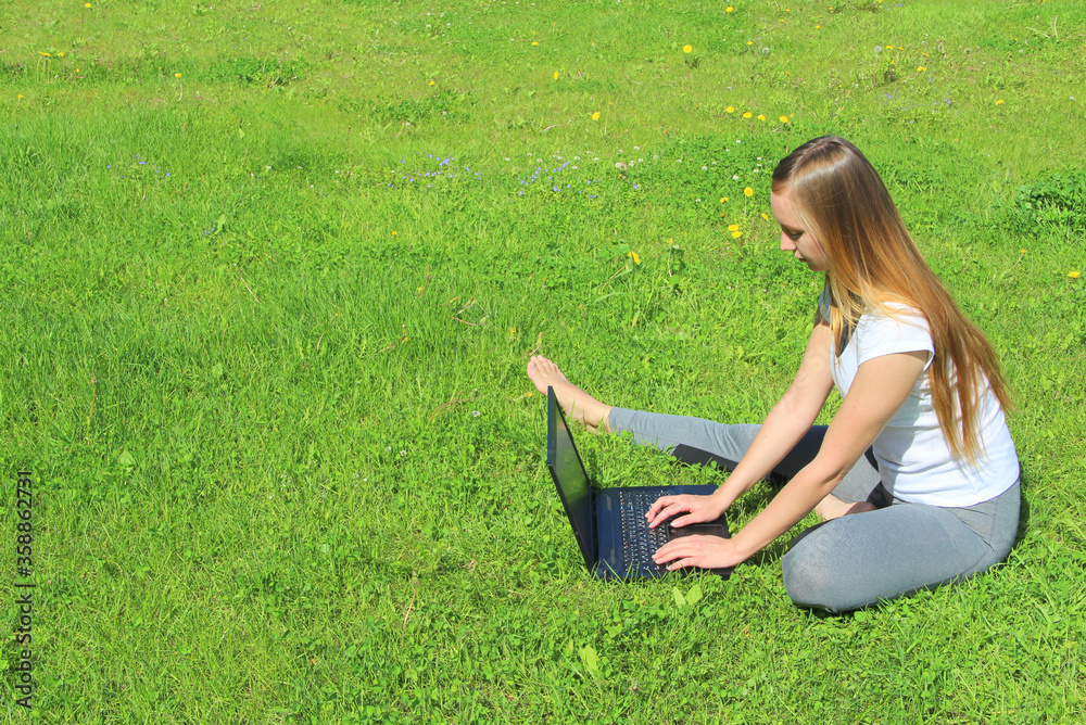 A beautiful young white girl in a white t-shirt and with long hair sitting on green grass, on the lawn and working behind a black laptop. A beautiful young white girl in a white t-shirt and with long hair sitting on green grass, on the lawn and working behind a black laptop.
