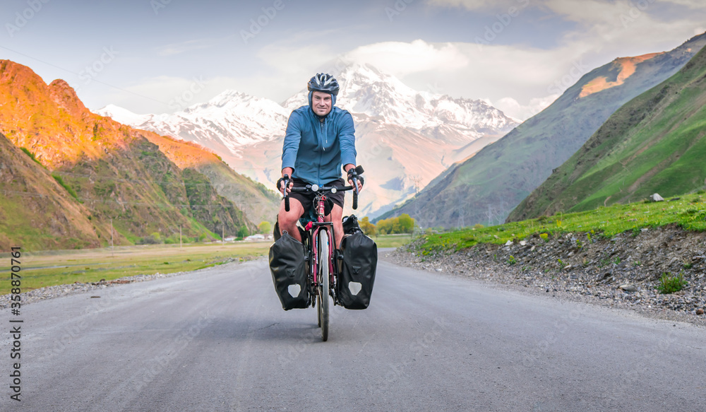 Male cyclist is on touring bicycle front side cycling bicycle touring with mountains white peaks background in summer or spring. Cycling holidays around caucasus in Georgia Male cyclist is on touring bicycle front side cycling bicycle touring with mountains white peaks background in summer or spring. Cycling holidays around caucasus in Georgia
