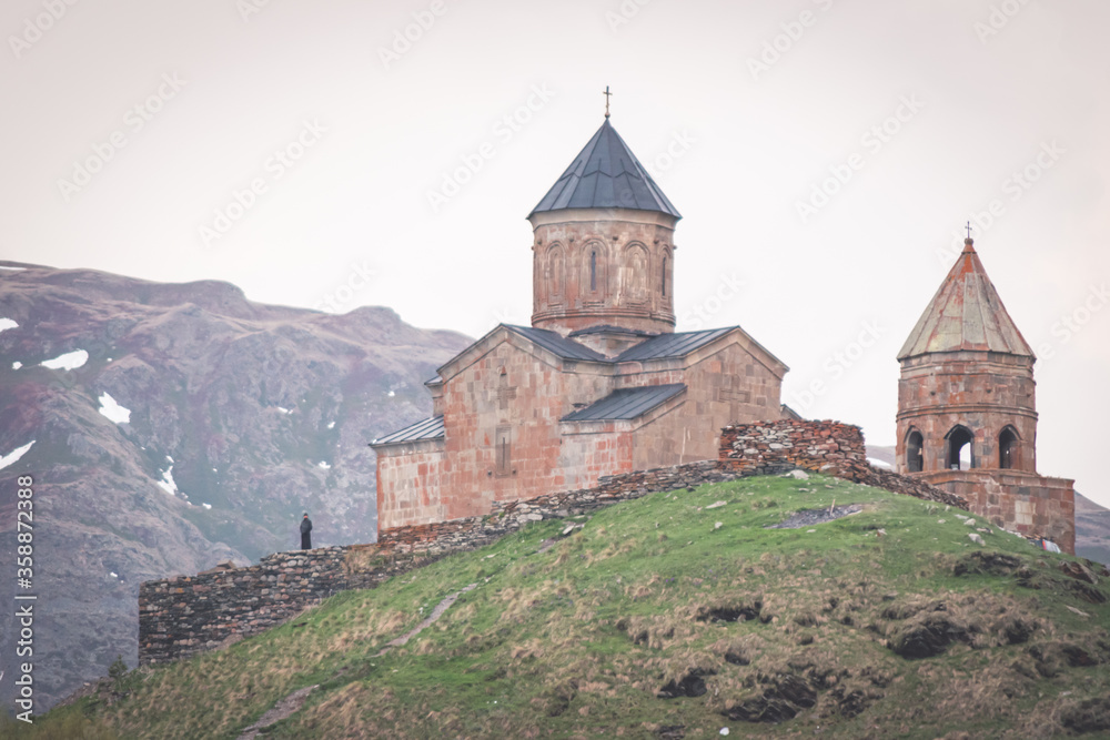 Gergeti trinity church on the hill with priest standing in black clothes nearby and enjoying panorama. Religion and saint places in Georgia. 2020