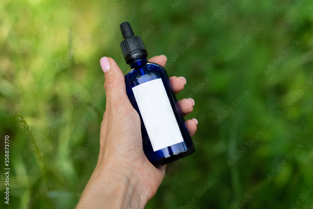 Fototapeta premium Against the background of green grass, a female hand holds a blue cosmetic bottle