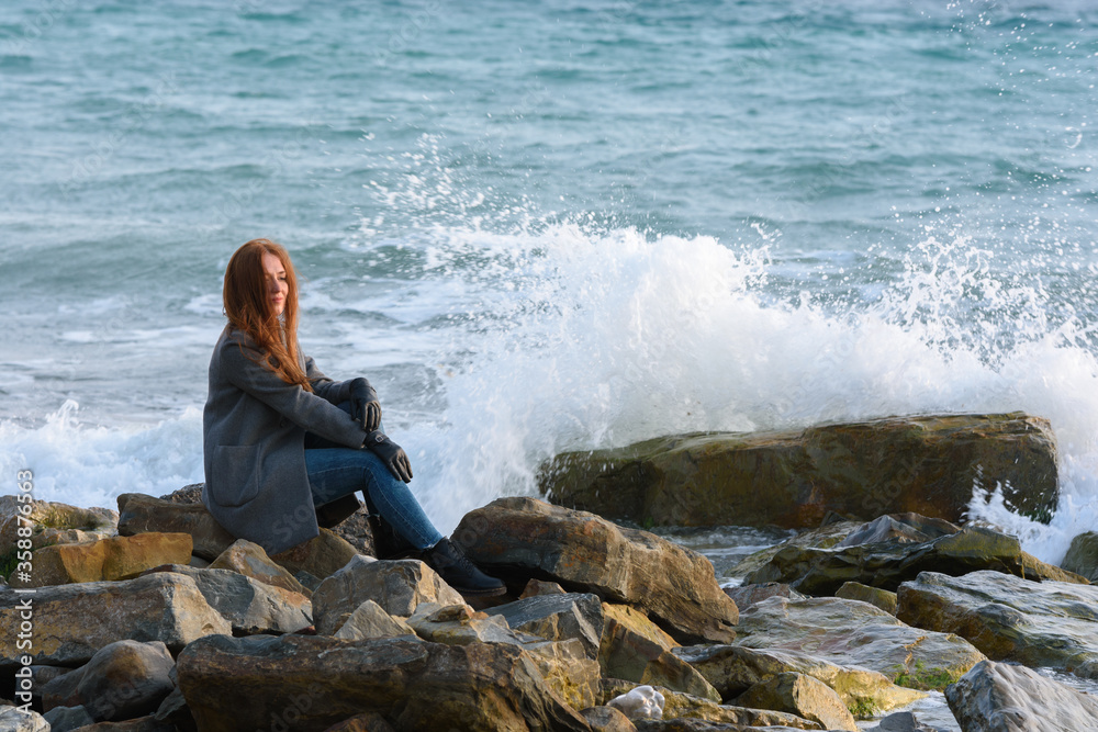 Girl in autumn windy weather sits by the sea on a rocky shore