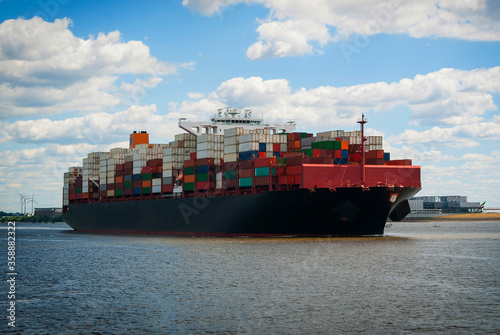 A cargo ship with many colorful containers floats along the river on a sunny day.