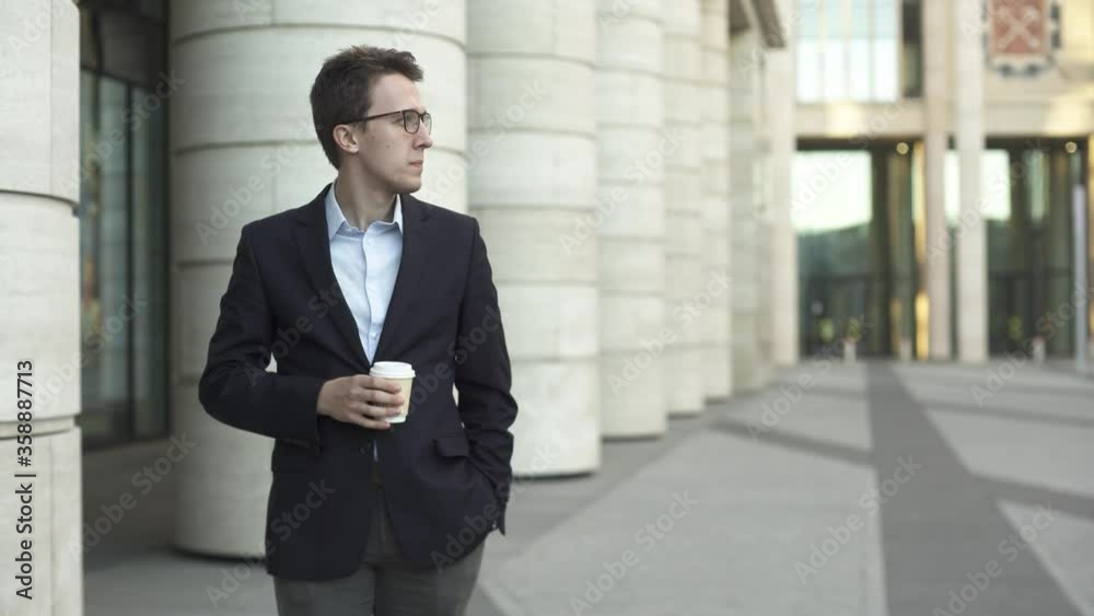 Young businessman in dark blue suit and glasses stand on the street near modern office building and drink coffee during his refreshment. Young businessman in dark blue suit and glasses stand on the street near modern office building and drink coffee during his refreshment.