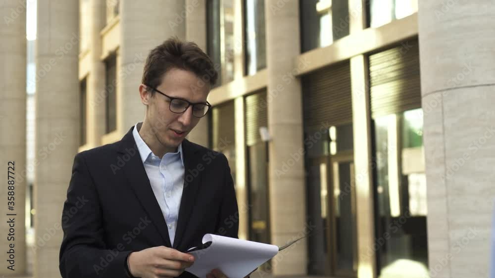Young businessman in dark blue suit and glasses checks documents while talking with his employee on the street near modern office building. Young businessman in dark blue suit and glasses checks documents while talking with his employee on the street near modern office building.