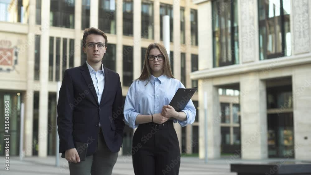 Young businessman in dark blue suit and businesswoman in formal suit are standing and looking in the camera near modern office building.