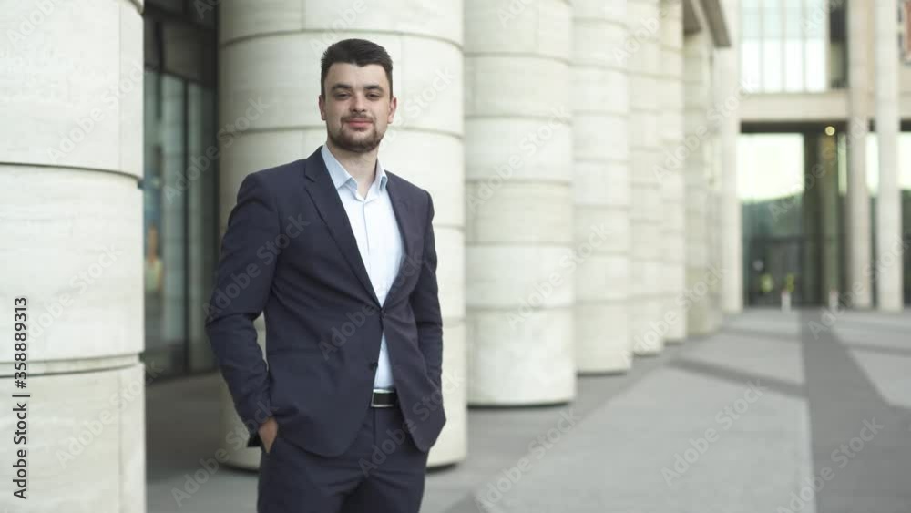 Young businessman in dark blue suit is standing and looking in the camera on the street near modern office building. Young businessman in dark blue suit is standing and looking in the camera on the street near modern office building.