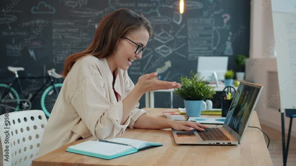 Happy young lady is talking to friends online with laptop making internet video call waving hand sitting at desk in office. Communication and technology concept.
