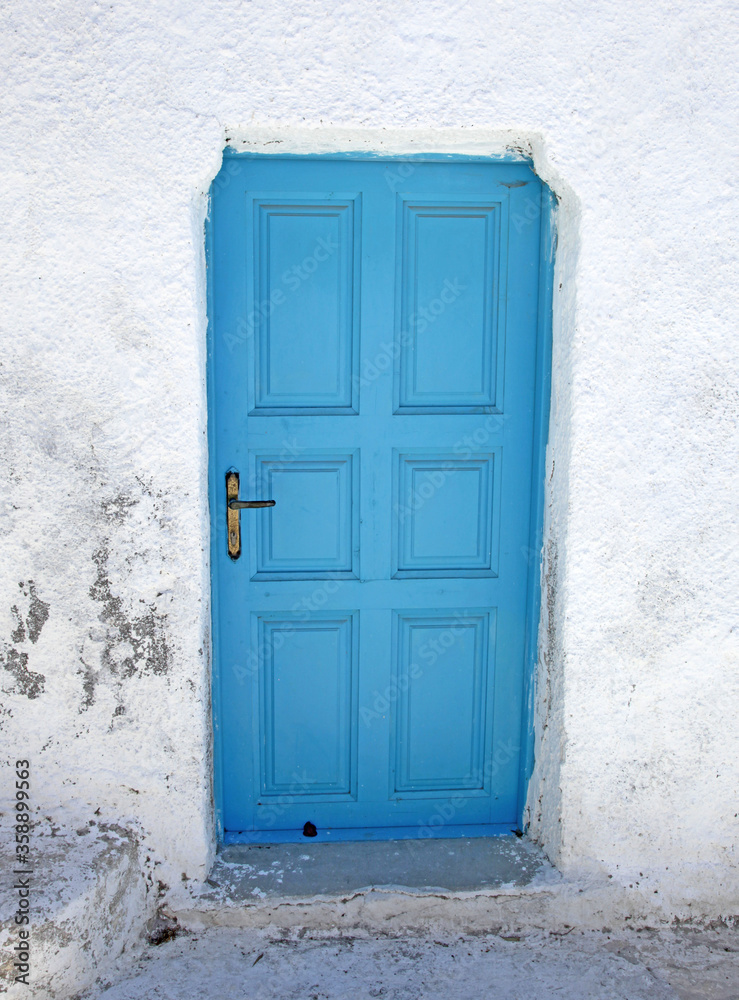A blue entrance door in a white wall on a street in the traditional village of Megalochori in Santorini, Greece.