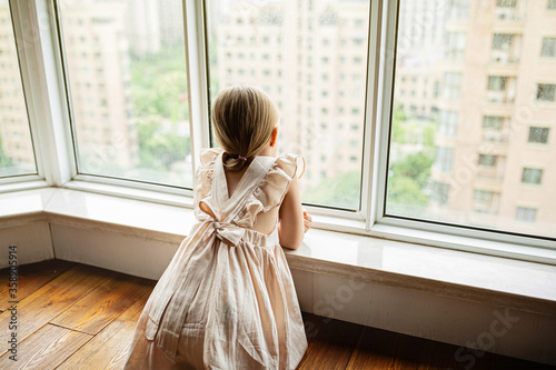 Fotografie, Obraz Stylish little girl with blonde hair sitting at home near window during coronavirus covid-19 self isolation