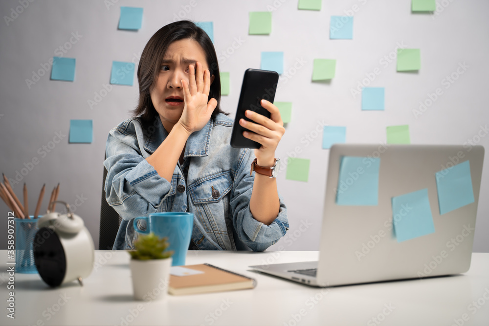 Asian woman shocked looking at smart phone reading news and screaming at home office. WFH. Work from home. Prevention Coronavirus COVID-19 concept. Asian woman shocked looking at smart phone reading news and screaming at home office. WFH. Work from home. Prevention Coronavirus COVID-19 concept.