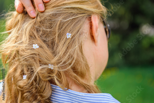 Back view of a woman hair with small white flowers on green blurred background. Blonde touches her curls, close-up. Natural hair care products, strengthening the hair roots decoction of herbs.