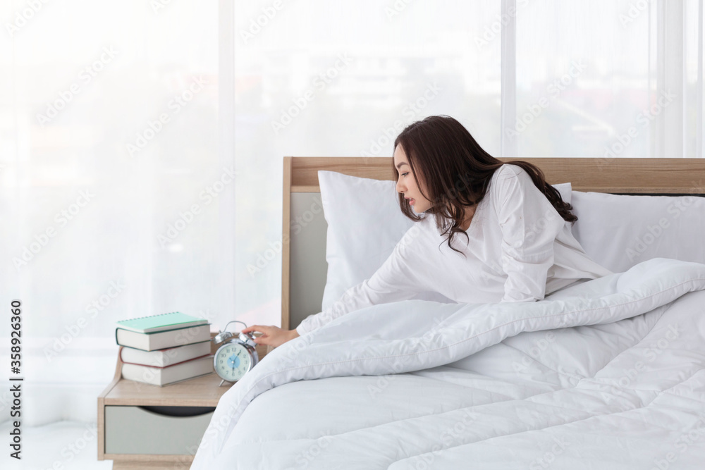 Woman in bed trying to wake up with alarm clock. Young girl in white on bed. Portrait of beautiful young asian girl with alarm clock in hand feeling frustrated on bed after getting up in morning. Woman in bed trying to wake up with alarm clock. Young girl in white on bed. Portrait of beautiful young asian girl with alarm clock in hand feeling frustrated on bed after getting up in morning.