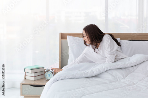 Woman in bed trying to wake up with alarm clock. Young girl in white on bed. Portrait of beautiful young asian girl with alarm clock in hand feeling frustrated on bed after getting up in morning.