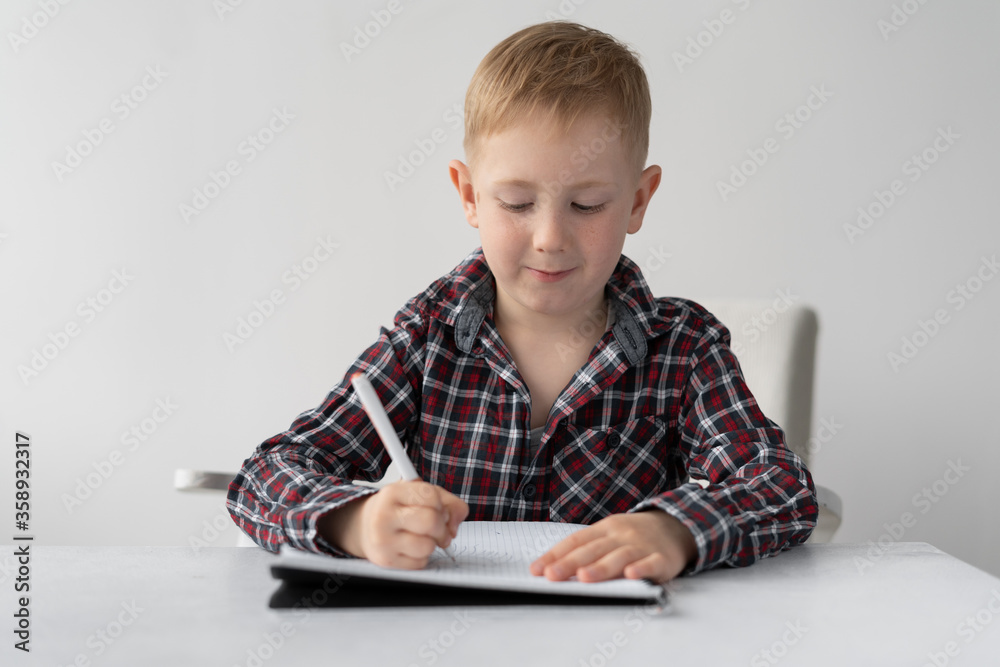A teenager studies remotely. The boy writes a task in a notebook with a pen. He sits at a table and listens to an online lesson on a webcam. Study and quarantine. A teenager studies remotely. The boy writes a task in a notebook with a pen. He sits at a table and listens to an online lesson on a webcam. Study and quarantine.