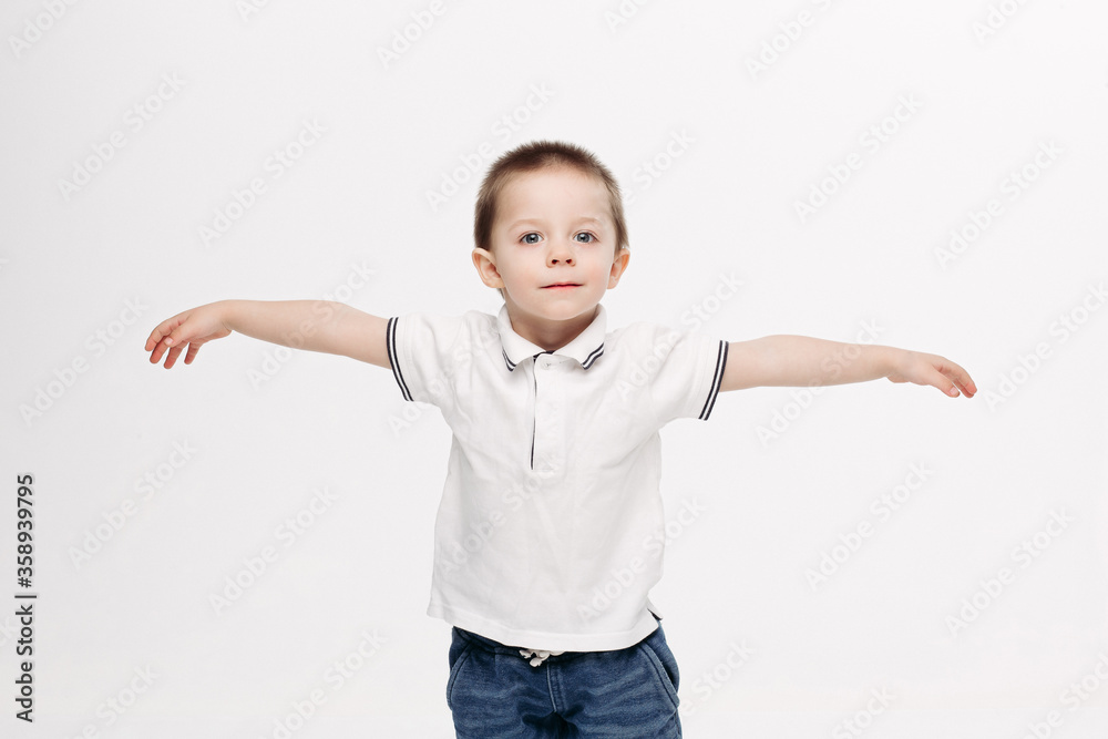Funny little boy with blue eyes, wearing in white polo t-shirt, jeans, gesturing by hands and showing wings like flying bird, in different sides. Kid playing at studio and enjoying. White background. Funny little boy with blue eyes, wearing in white polo t-shirt, jeans, gesturing by hands and showing wings like flying bird, in different sides. Kid playing at studio and enjoying. White background.
