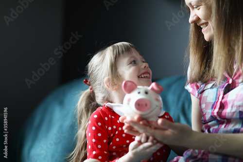 Portrait of smiling attractive funny toddler sitting on couch indoors. Cute kid saving up for future in piggy bank. Mum and little girl spending time together. Childhood and parenthood concept photo