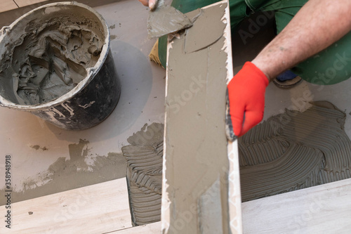 The builder applies cement adhesive to the ceramic tile with a stainless trowel.