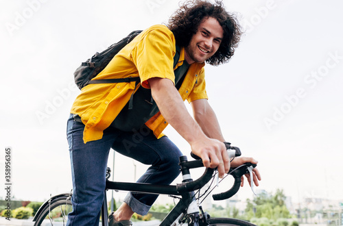 Happy man cycling on his bike in the city street. Cheerful male courier with curly hair in yellow shirt delivers parcel cycling with a bicycle in the city.