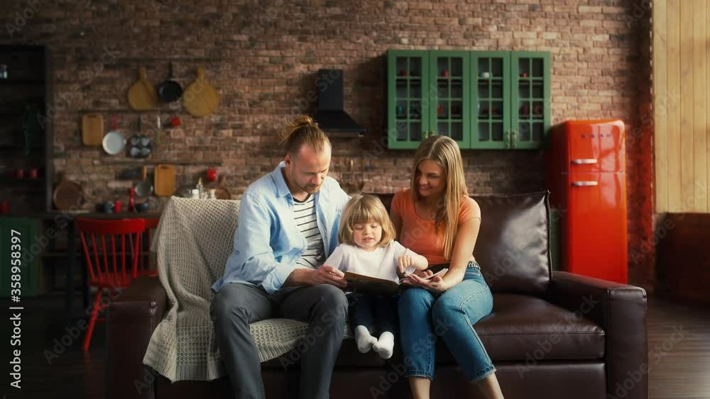 Couple and child boy are sitting on couch in studio apartment. Little one playing with kids magazine and parents looking at him. Slow motion Couple and child boy are sitting on couch in studio apartment. Little one playing with kids magazine and parents looking at him. Slow motion