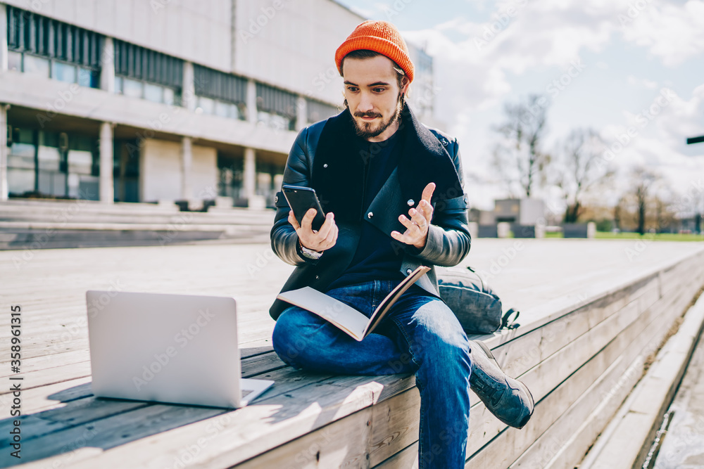 Confused Caucasian male student puzzled with received web information with exams schedule, millennial hipster guy dressed in trendy clothing reading message during e learning at urban setting