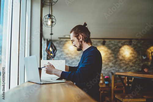 Side view of serious skilled male student studying information for knowledge textbook for put into chapter of course work, millennial man analyzing data from laptop comture and paper notepad