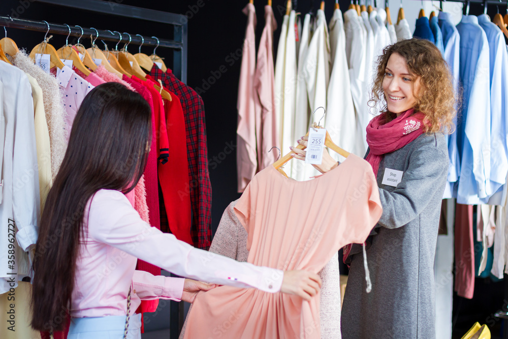 Young woman is choosing, buying fashionable dress in store, boutique in center, mall. Kind seller, consultant, stylist, shop assistant is serving girl. Black friday. Shopping and sale concept.