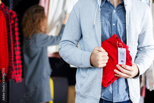Closeup of young man is stealing red jeans in store, shop, boutique at shopping center. Guy is hiding unpaid good under clothes. Seller, assistant caught thief on hot. Shoplifting concept.