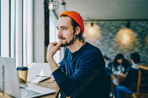 Contemplative hipster guy dressed in stylish apparel thoughtful looking away while listening interesting audio book via electronic headphones, pondering young man thinking about 4g internet for laptop photo